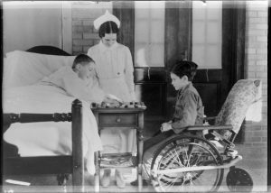 Grayscale wide shot of two children playing checkers with a nurse standing in the middle. The checkers board is placed on a wooden end table with a drawer. A child on the right sits in a wheelchair and gazes at the board. A child on the left smiles as they move a peon while lying on a bed.