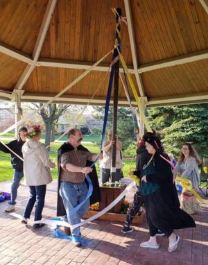 Milwaukee's pagan community members perform a Maypole dance in a gazebo during a Beltane celebration. They surround a pole while holding their own ribbon that is attached to the pole. Someone on the left wears a crown of flowers. The gazebo's ceiling and floor are visible. Trees and green lawns are seen in the background.