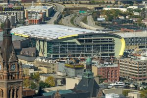 Bird's eye view of Fiserv Forum under construction. The iconic roof that is still being built appears from afar. Milwaukee's landscape surrounds the area. The City Hall's bell tower and steeple are visible in the foreground; the highway and its entrance are visible in the background.