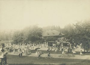 Sepia-colored elevated view of a crowd traversing a pathway at Washington Park. Most of them walk, some leading their bicycles to the right. A building is visible in the far background between lush tall trees. Two horse-drawn carriages with the "Bitkers" logo on their sides appear among the crowd in the image's center and center-right. A young boy in the left foreground stands nearest and faces the camera lens.