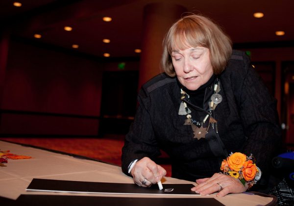 Medium shot of Betty Quadracci from the waist up, bending towards the camera lens while signing a signature plaque with her right hand. She wears a black long-sleeve blouse, a necklace, and a wrist corsage on her left hand. Her eyes look at the plaque on a long table covered with a white tablecloth. A hall with dim light and a red carpet are in the background.