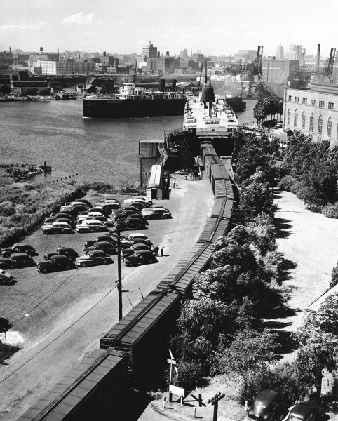 An aerial photo shows the City of Saginaw car ferry that is docked and being loaded with a long line of railway cars. Dozens of cars parking next to the railroad seem to indicate a busy day.