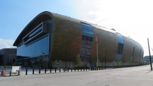 Long-shot of the Fiserv Forum exterior showcases its front and side view and the swooping roof. The Milwaukee Bucks monument sign appears next to the facade. A concrete pedestrian area is visible in the foreground.