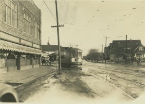 Sepia-colored long shot of an electric street car in the image's center back running towards the foreground. The trolley tracks and the muddy street are visible in the foreground. An automobile appears behind the streetcar. Overhead wires and utility poles can be seen. Buildings can be seen in the left foreground and along the street in the background.