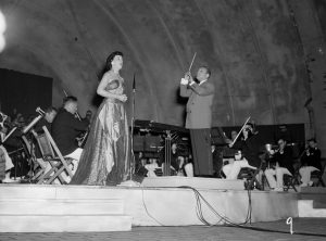A woman in a long dress faces to the right while singing with an orchestra playing behind her in Washington Park's bandshell. The singer and the orchestra conductor in a suit stand in the image's center. The stage, cables, and the bandshell's arched wall are visible.