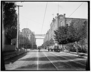 Sepia-colored long shot of one entrance of the Pabst Brewery complex. The image shows a long road stretching down in the center. A welcoming gateway that reads "Pabst" is above the street in the distance. The grand structure of the brewery sits on the right along the street in the background. People and horse-drawn wagons appear on the road.