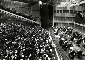 High-angle shot of a side view of a theater's stage where members of the Bel Canto Chorus perform with the Milwaukee Symphony Orchestra. The musicians with instruments perform on stage while hundreds of singers stand in the seating areas.