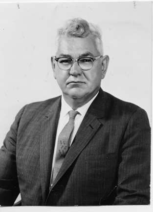 Grayscale medium shot of Harold Breier in glasses and notched lapel suit. He poses in an upright body posture and direct eye contact with the camera lens showing an expression of confidence.