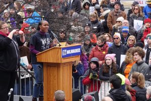 Bria Smith speaks on a podium with a sign reading "March For Our Lives Milwaukee" on the left, standing next to a sign language interpreter. Both are facing right to the crowds. A large group of people stands below, watching Smith in their warm clothes. Some hold protest signs.