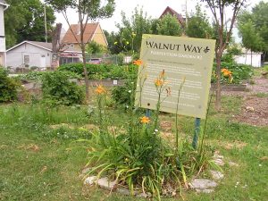 A wide shot of Walnut Way Community Garden displays a board that reads "Walnut Way Production Garden #2." It is installed among green grass and flowers. Trees, raised garden beds filled with plants, and roofs of houses in the neighborhood are visible in the background.