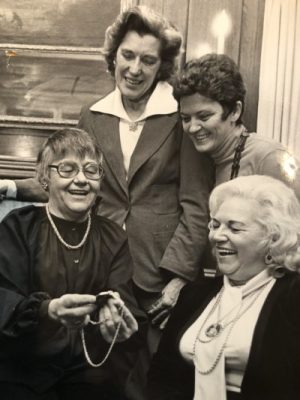 Catherine Conroy sits on the left in glasses and long sleeved blouse showing a necklace she holds with both hands to other women beside her. Their eyes look at the chain while smiling in their formal attire.