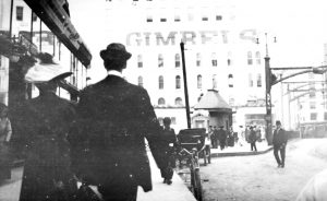Rear view of two people walking on a sidewalk towards Gimbels Department Store. The person on the left wears a dress and a large hat while the other is in a suit and a smaller round hat. A group of people crowd the entrance of the store. A big "Gimbel’s" sign appears on the building's exterior wall.