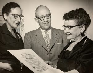 Sepia-colored medium shot of three people in formal attire smiling while standing next to each other. The one on the right is Charlotte Partridge in glasses carrying a piece of paper. The one on the left is Miriam Frink with eyes looking at the document. A man in a suit and tie stands in the middle while glancing at Partridge.