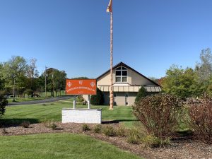 Long shot of the gurdwara building in Brookfield. The place's orange-colored monument sign appears in its yard. Several plants and green lawns embellish the yard in the foreground. Tall green trees are visible in the background. Above is the clear blue sky.