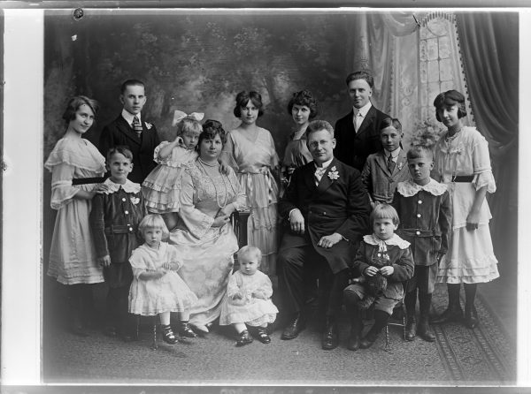 Grayscale family portrait of the Uszlers and their 13 children in dresses and suits. Louis E. and Wanda Uszler and three small children sit in the middle. Older children stand side by side surrounding their parents. Everyone's clothes are elegant. Wanda Uszler wears a pearl necklace and Louis Uszler wears a boutonniere.