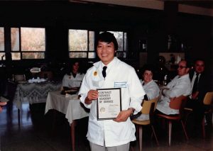 Medium shot of Nao Shoua Xiong smiling in white clothes while showing a certificate that reads "Contract Services Academy of Services." Behind Xiong, who poses in a standing position, is a group of people sitting at a dining table.