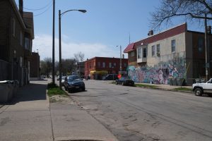 Long shot of a street stretching down at the image center. Clarke Square Mural appears in the distant right. It is painted on the lower part of a wall of a two-story building next to a road junction. Cars park here and there on the street side. Visible in the left foreground are a building's side view and a sidewalk.