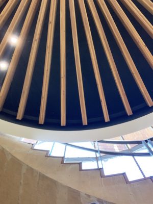The image's upper portion shows a set of wooden-like structure that composes the underside of a central oculus light at the Indian Community School. The image's bottom displays the side part of a stairway.