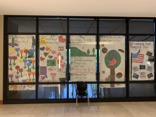 Five sets of colorful paper crafts are hung on the interior windows of a classroom. Each sheet of newsprint carries words written in Anishinaabemowin and English as well as numbers corresponding to various days in May. The first from left is entitled "Ma Hitawus Haap! May Day!" decorated predominantly with flower-shaped cutouts. The second has heart-shaped papers and is entitled "Hi'uni haap! Mother's Day!" The third sheet contains a list of May holidays. The fourth is themed National Apple Pie Day and the last one represents Memorial Day.